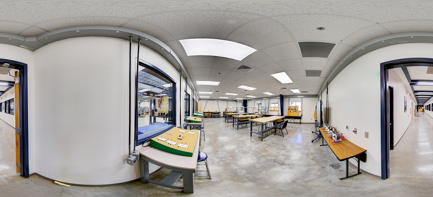 Panoramic view of an electronics or technical lab area with worktables, small equipment setups, and instructional stations, connected to hallways and adjacent rooms with large interior windows.