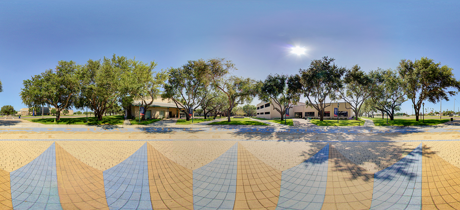 Panoramic outdoor view of a campus walkway lined with trees casting shadows, with low academic buildings on either side, patterned paving in the foreground, and a bright sun overhead in a clear sky.