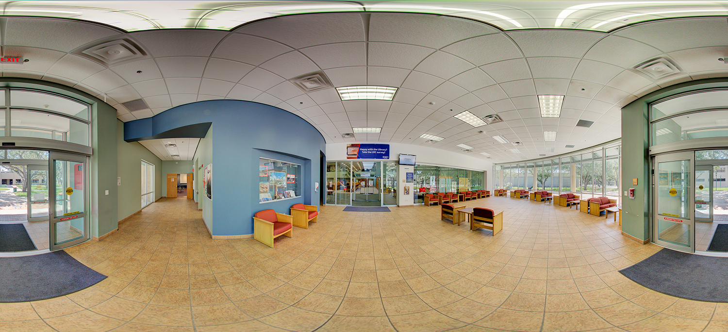 Panoramic view of a campus lobby with tiled floors, cushioned seating areas, and large glass windows, featuring entrances on both sides and a central doorway leading into a library or common area.