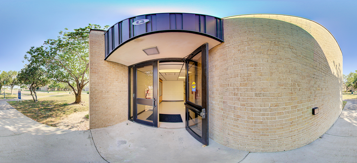 Panoramic view of a curved brick building entrance with glass double doors open to a hallway, shaded by a small overhang, with trees and walkways visible outside on a sunny day.