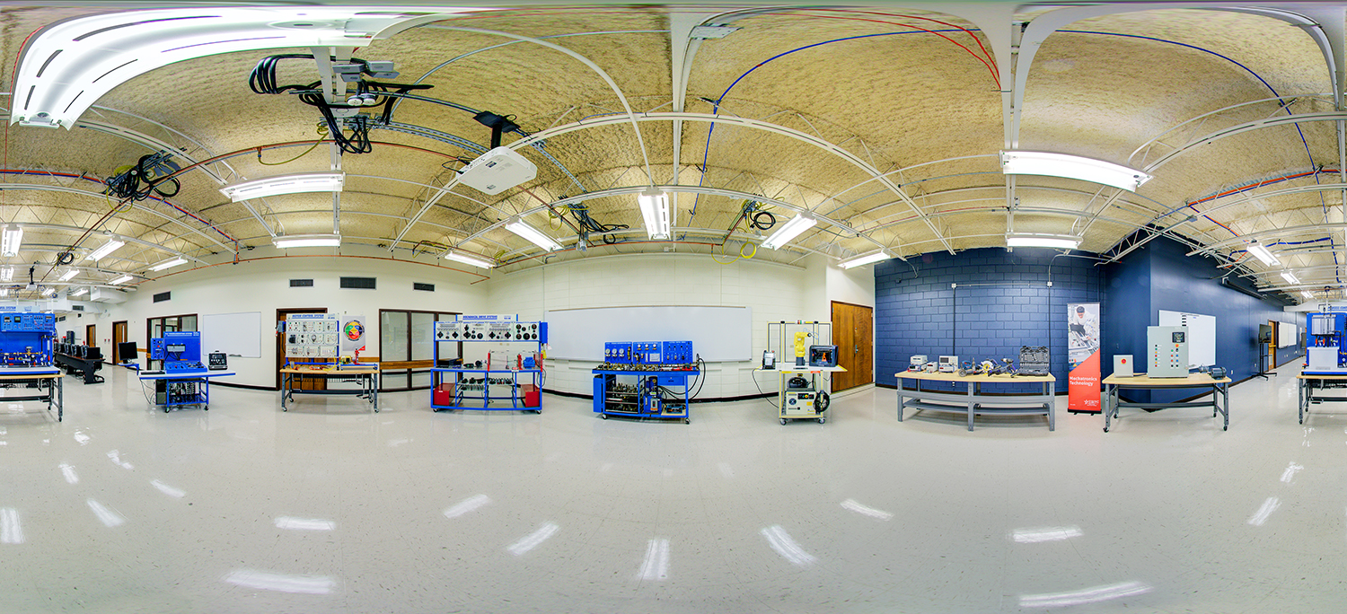 Panoramic view of a bright technical training lab with polished floors, exposed ceiling wiring and lights, and multiple workstations equipped with blue industrial training equipment and control panels arranged along the walls.