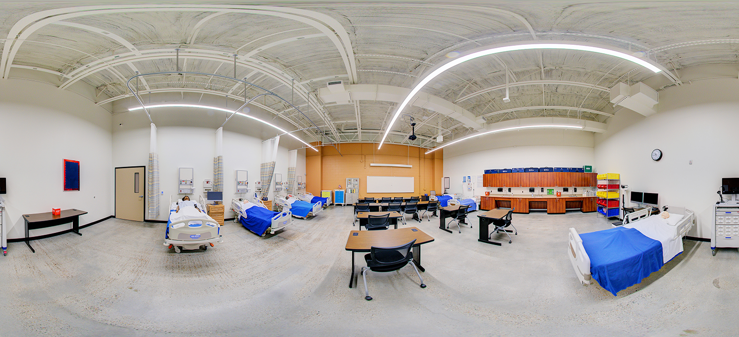 Panoramic view of a nursing skills lab with multiple hospital beds, patient mannequins, and medical equipment arranged along the walls, with classroom tables and chairs in the center and storage cabinets along the back wall.