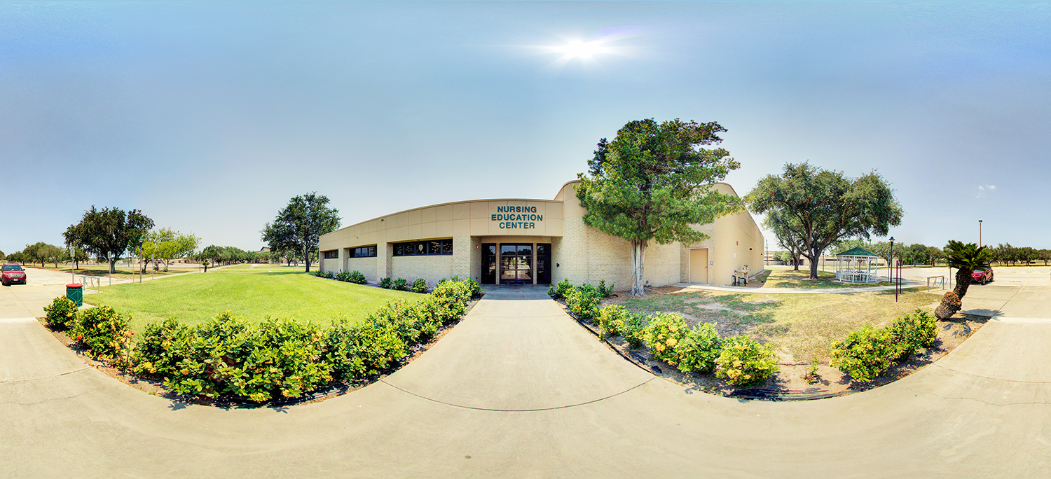 Panoramic view of a single-story campus building labeled “Nursing Education Center,” with a curved facade, central glass entrance, surrounding green lawn, shrubs, and trees under a bright sunny sky.