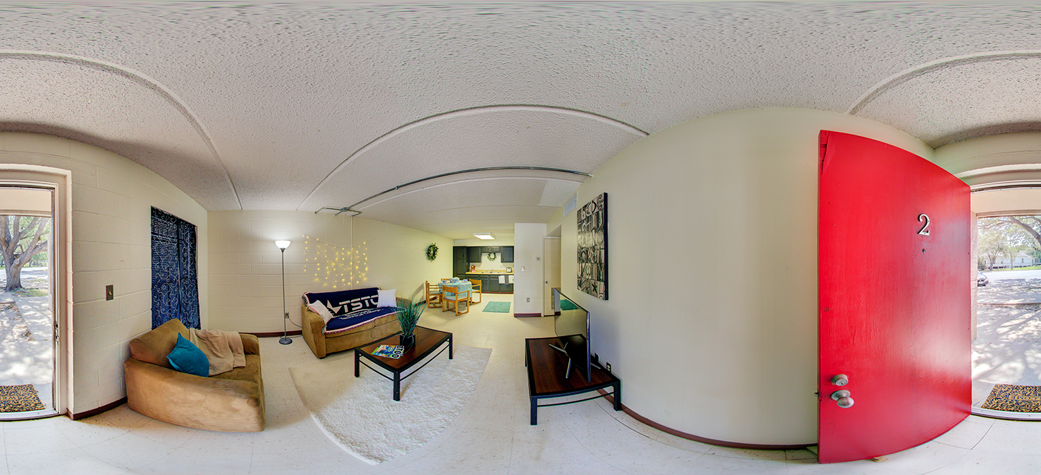 Panoramic view of a small apartment-style living space with a red front door, a cozy seating area with couches and a coffee table, a TV on a stand, decorative lighting, and a kitchenette with a dining table in the background.