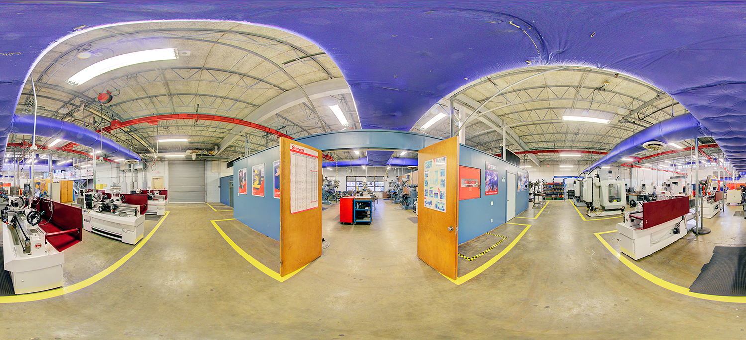 Panoramic view of a machine shop lab with lathes, milling machines, and workstations arranged around a central enclosed classroom area with open double doors, under exposed ductwork and overhead cranes.
