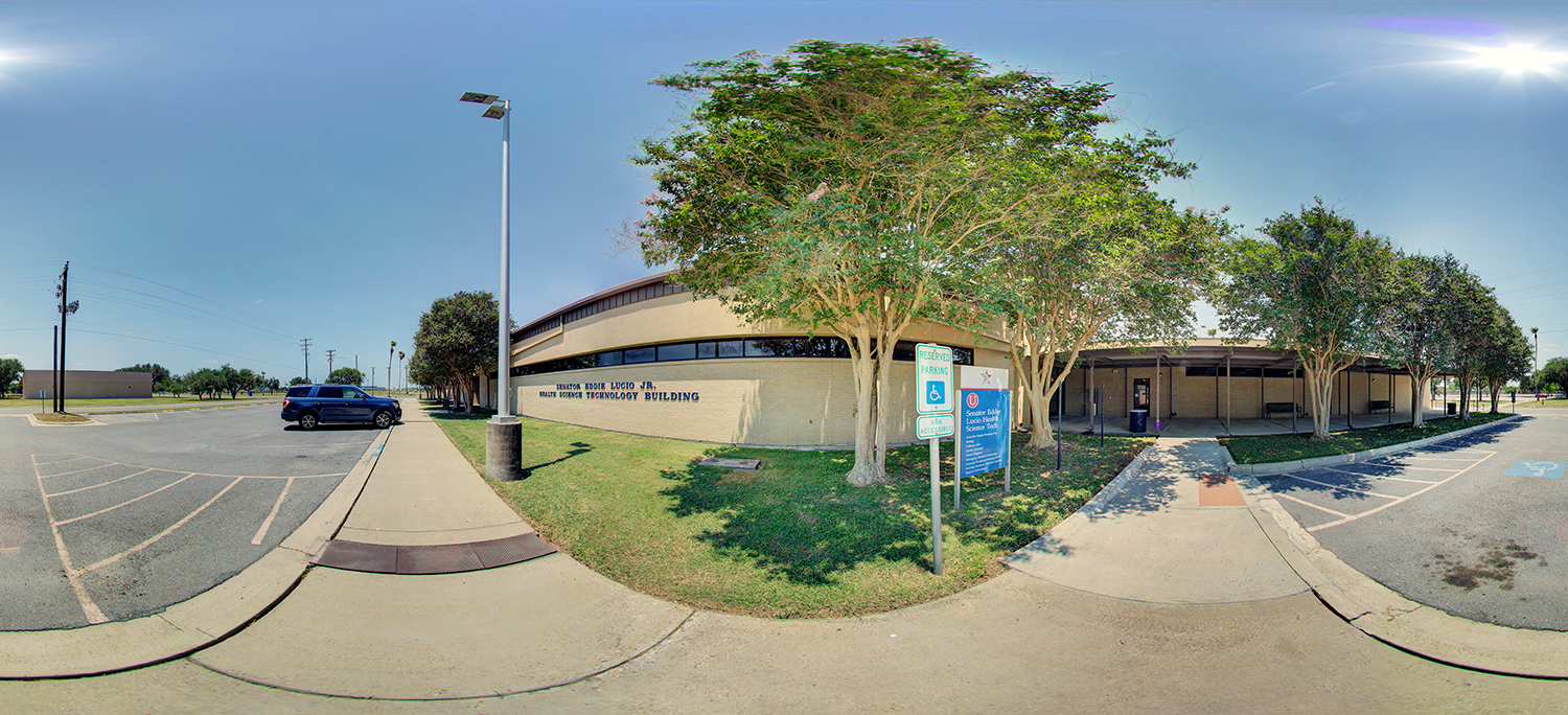 Panoramic view of a campus building labeled “Health Science Technology Building,” with a shaded entrance, nearby accessible parking signs, trees lining the walkway, and a mostly empty parking lot under a bright sunny sky.