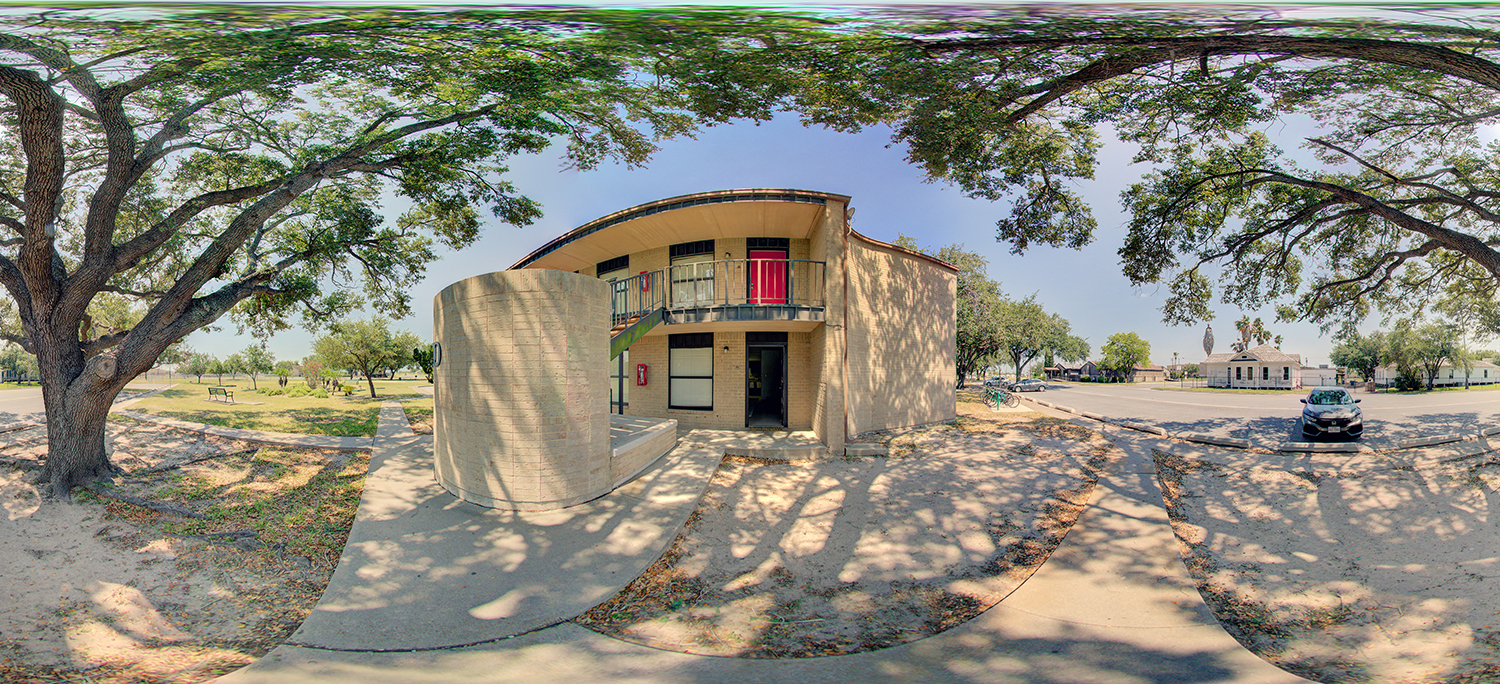 Panoramic view of a two-story campus building with a curved facade, exterior staircase, and red doors on the upper level, surrounded by trees, walkways, and a nearby parking area under bright daylight.