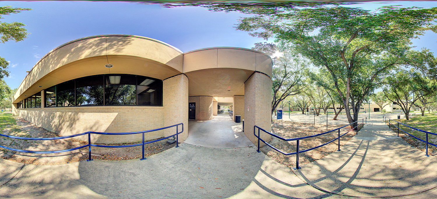 Panoramic exterior view of a curved, beige brick building with large tinted windows, a covered walkway entrance, and blue handrails lining accessible ramps, surrounded by trees casting dappled shade on the ground.