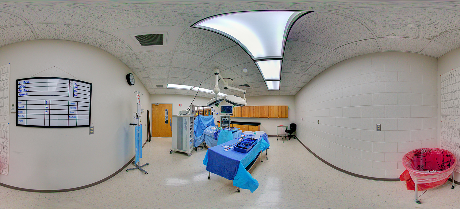 Panoramic view of a medical simulation room set up like an operating theater, featuring a patient mannequin on a surgical table, overhead surgical lights, instrument trays, medical equipment carts, and cabinets along the wall under bright clinical lighting.
