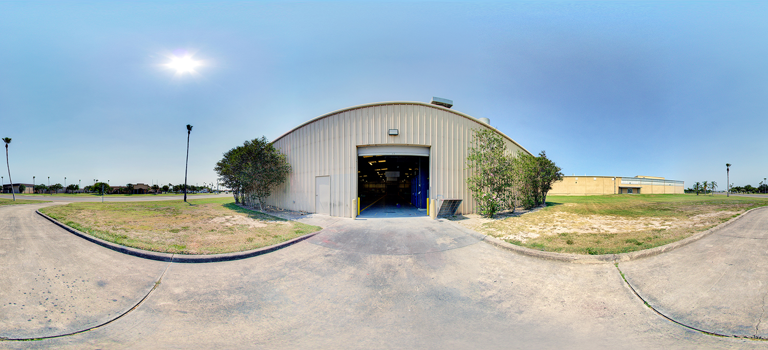 Panoramic exterior view of a large metal warehouse-style building with an open garage door, surrounded by grass, sidewalks, and a paved drive under a bright, clear sky with the sun overhead.