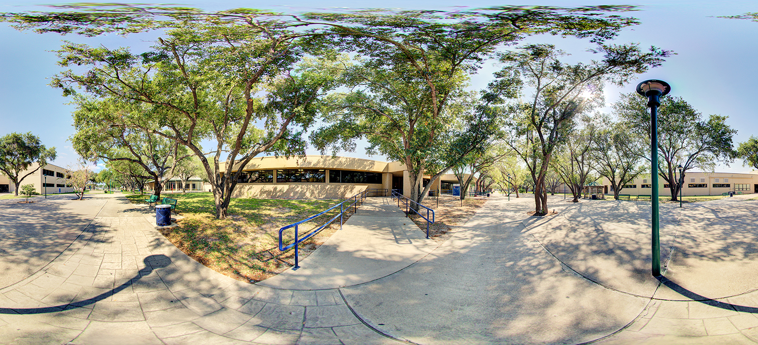 Panoramic view of an outdoor campus area with walkways, trees providing shade, a low building with large windows, blue handrails along a ramp, and open paved spaces with benches and a lamppost.