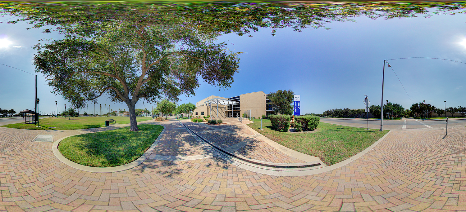 Panoramic view of a campus entrance with a large shade tree beside a curved brick walkway leading to a modern building with glass doors, surrounded by trimmed grass, shrubs, and nearby streets under a bright, clear sky.