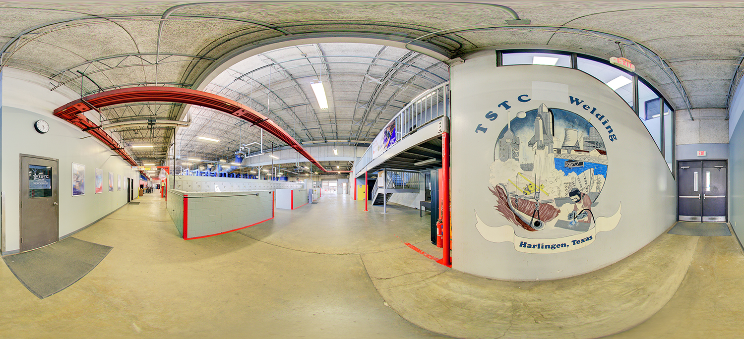 Panoramic view of a welding technology training facility with open lab bays, red overhead crane rails, and a large wall mural reading “TSTC Welding, Harlingen, Texas,” alongside industrial work areas and mezzanine storage.