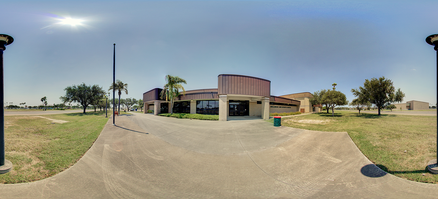 Panoramic view of a low, modern campus building labeled “Wellness and Sports Center,” with a curved facade, large windows, and a wide concrete walkway, surrounded by grass, scattered trees, and lamp posts under a bright sunny sky.