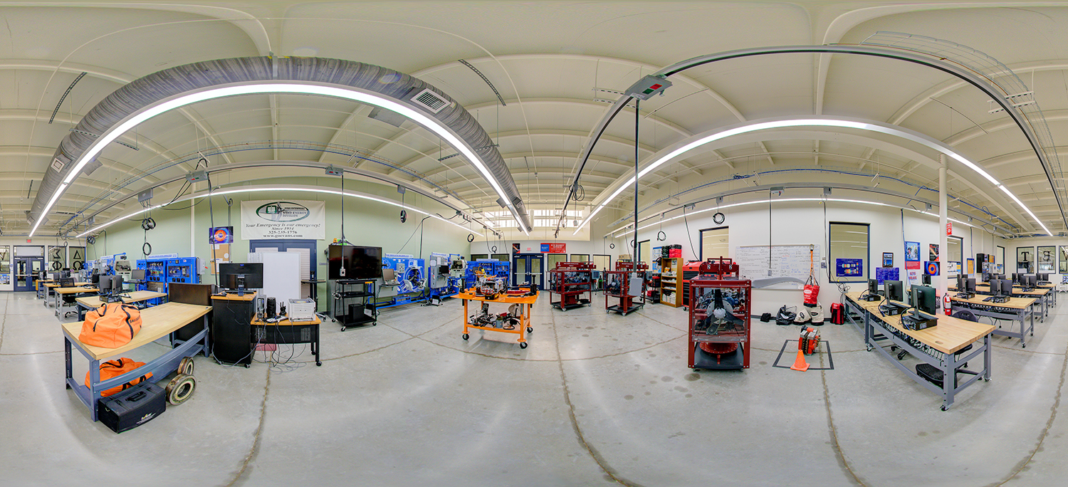 Panoramic view of an advanced technical lab area with workbenches, computers, and mechanical training equipment, including engine systems and industrial training rigs, under a high ceiling with exposed ductwork and lighting.