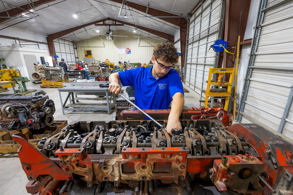 A student in safety glasses and a blue TSTC polo crouches while using a pneumatic impact wrench to loosen lug nuts on a large truck wheel inside a mechanic training workshop.