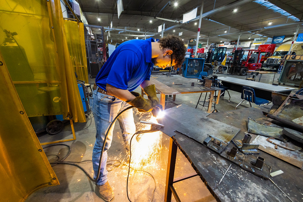 A student wearing safety glasses and gloves uses a cutting torch on a metal sheet, producing bright sparks while working at a table in a welding and fabrication lab.