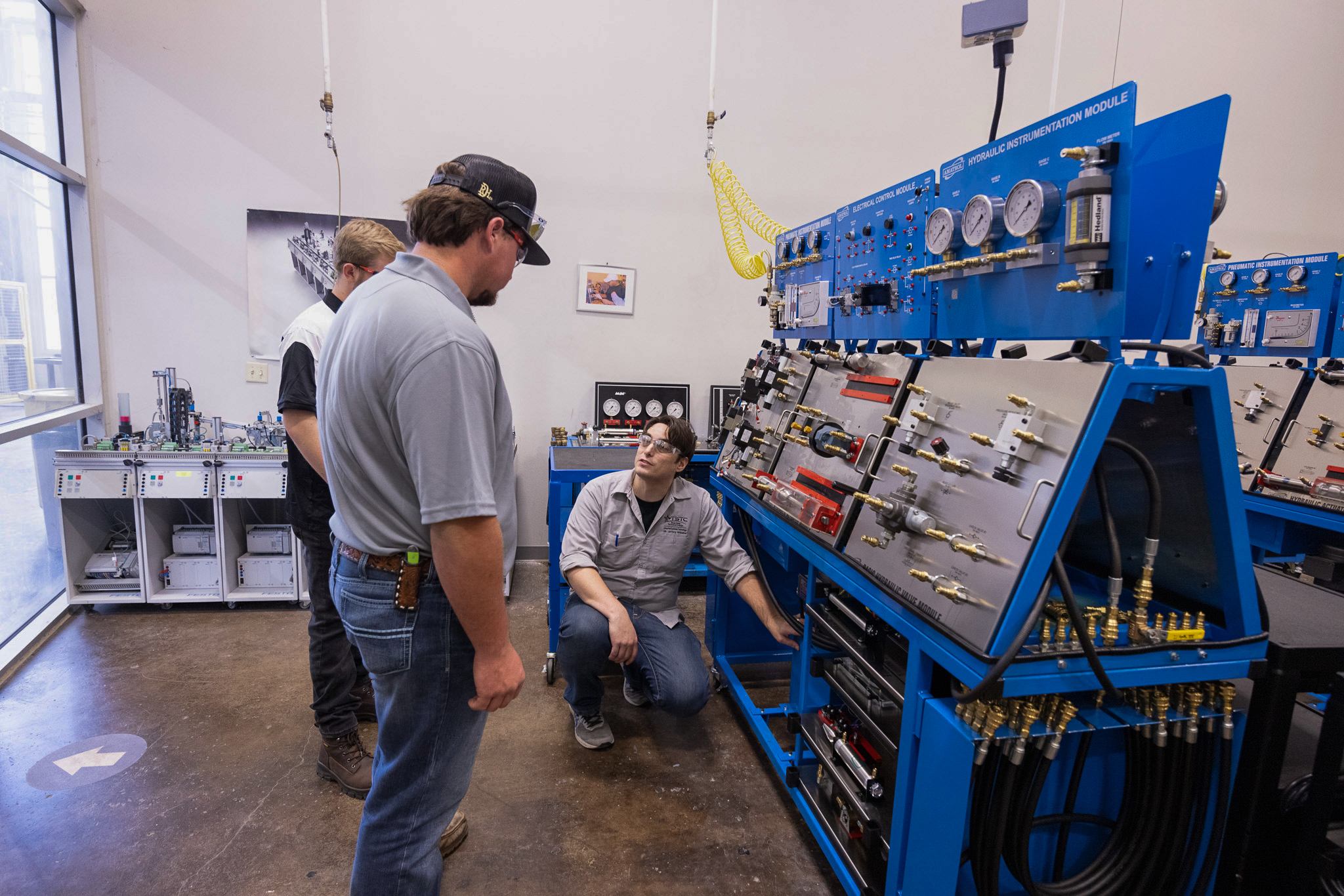 Three students in a lab gather around a blue hydraulic training system with gauges, valves, and hoses, as one kneels and explains components while the others observe.