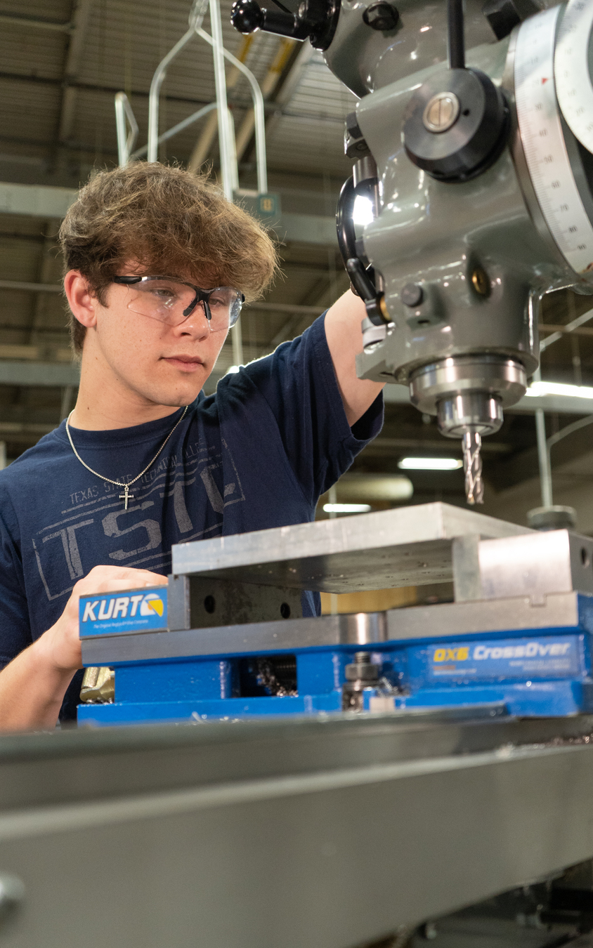 A student wearing safety glasses carefully lowers a drill bit from a milling machine toward a rectangular metal block clamped in a vise, focusing on precise positioning in a workshop.