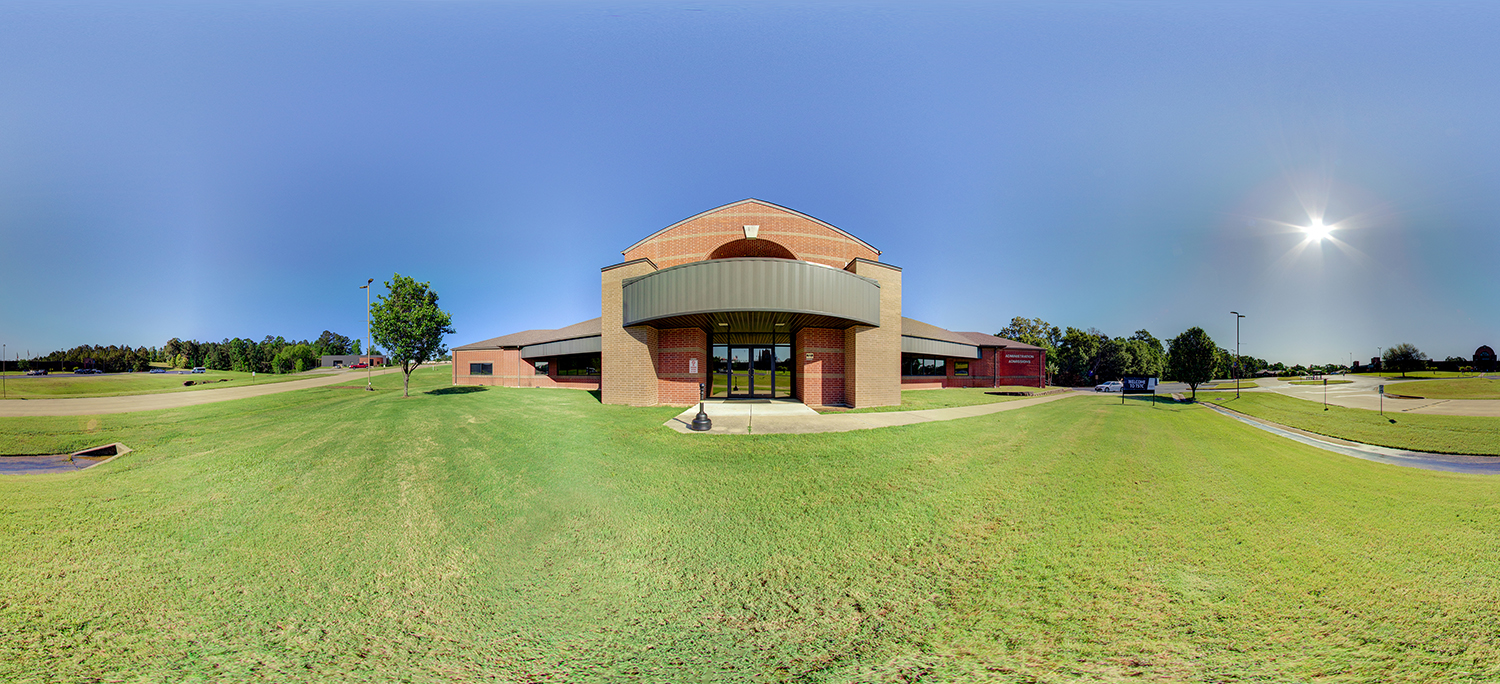 Panoramic exterior view of a low, brick academic building with a covered entrance, surrounded by green lawn, sidewalks, and a clear blue sky with bright sun overhead.
