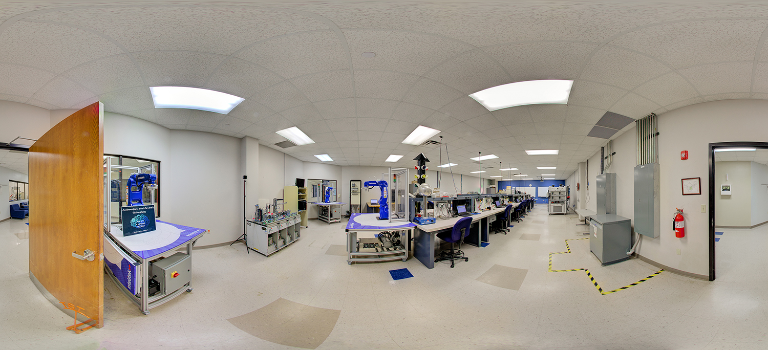 Panoramic view of a technical training lab equipped with robotics arms, control systems, and electronic workstations, where long benches with computers and instruments line the room, and instructional equipment is arranged for hands-on learning in a bright, organized space.