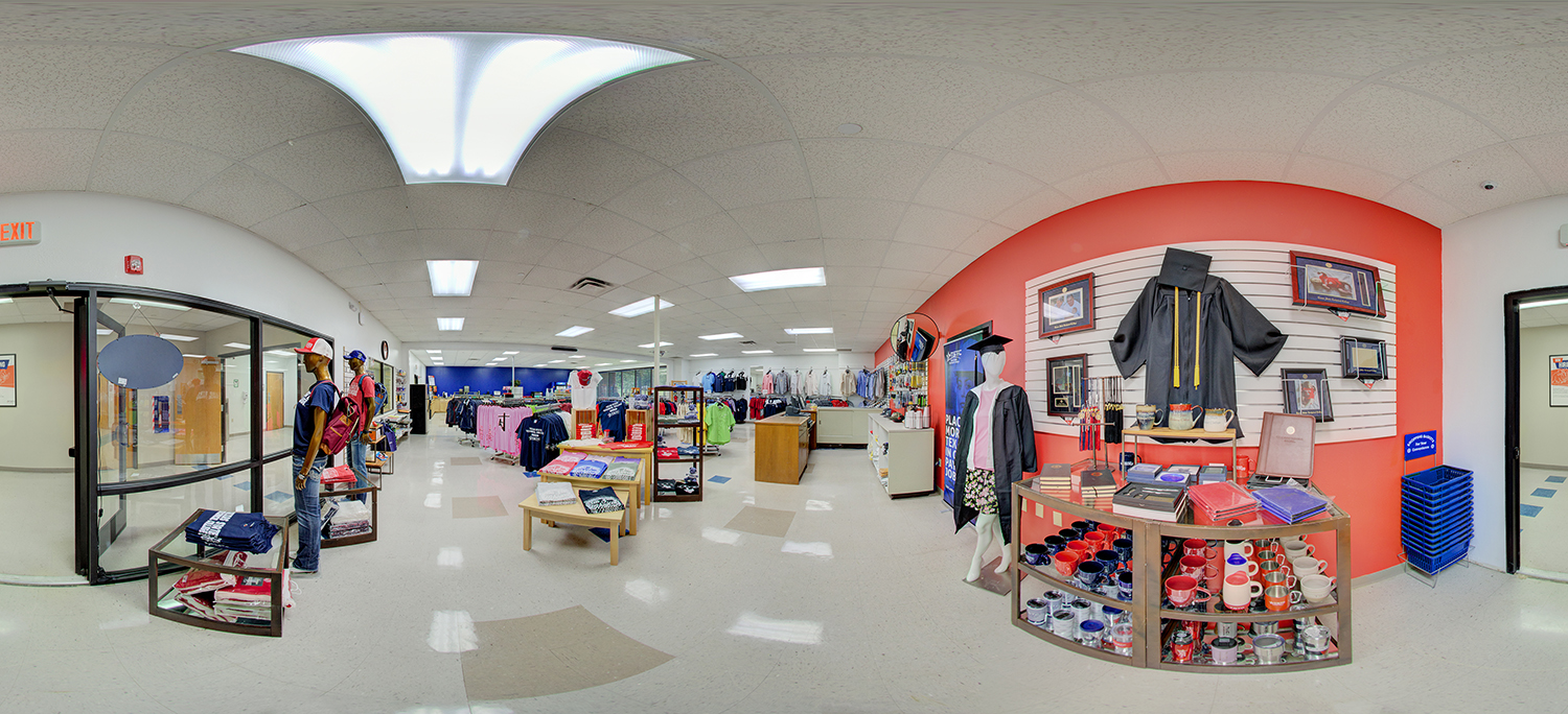 Panoramic view of a campus bookstore or retail space displaying school apparel, graduation items, and merchandise, with mannequins, clothing racks, and shelves of mugs and accessories against a red accent wall.