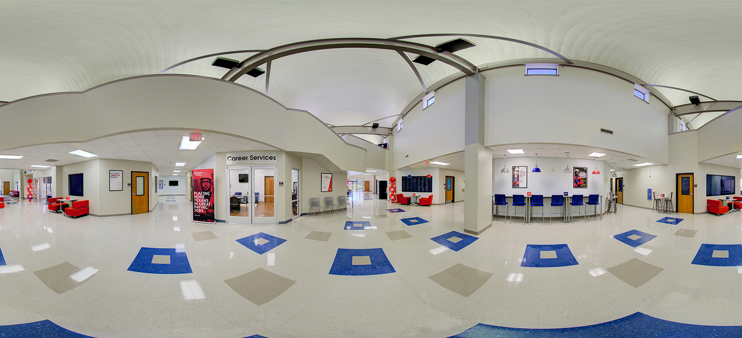 Panoramic view of a spacious campus lobby with a high curved ceiling, Career Services office, seating areas with red chairs, a row of blue stools along a counter, and open hallways leading to classrooms.