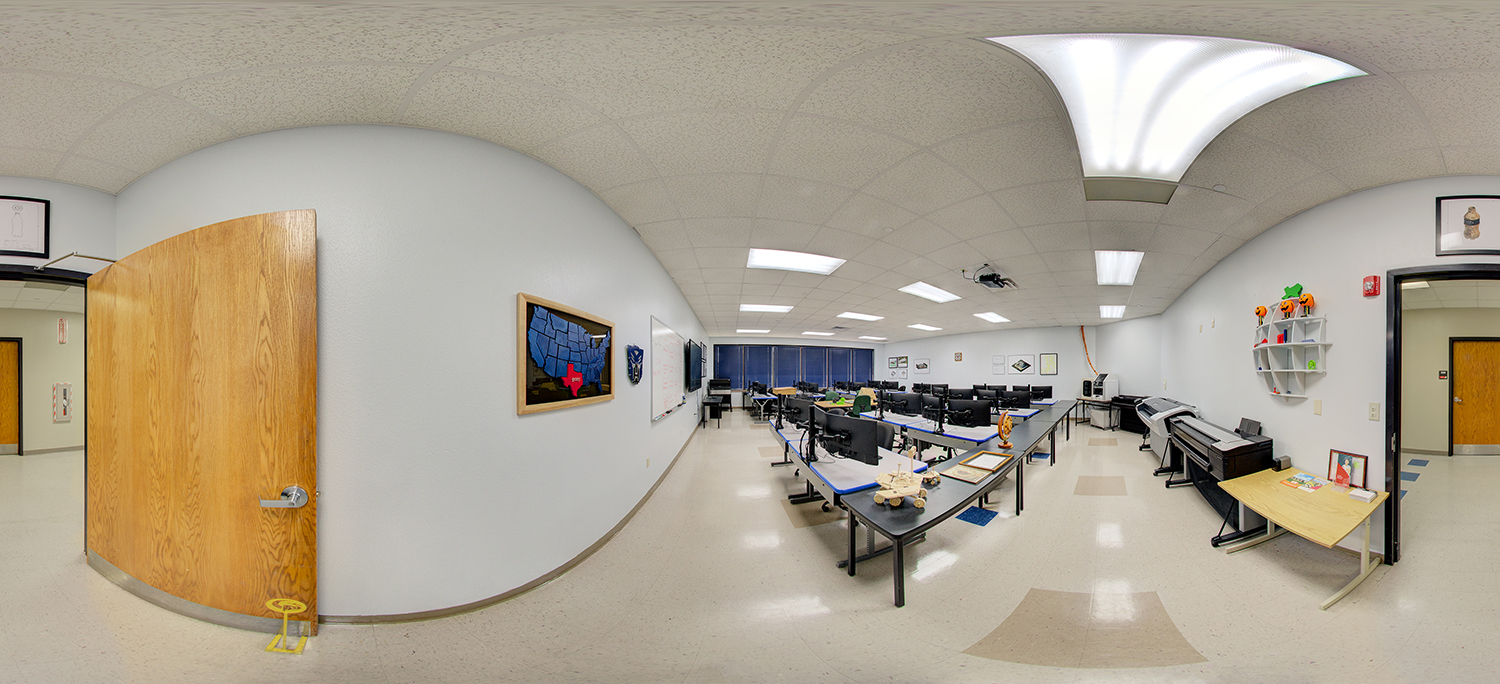 Panoramic view of a computer lab classroom with rows of desktop workstations on long tables, large-format printers along one wall, and instructional materials and posters displayed, all arranged in a clean, well-lit learning environment.