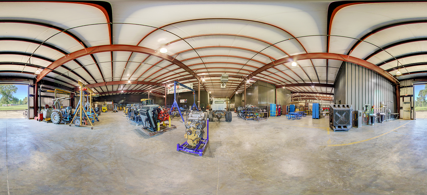 Panoramic view of a large industrial training workshop with high ceilings, exposed steel beams, and multiple heavy-duty engines, tractors, and mechanical equipment arranged across a spacious concrete floor.