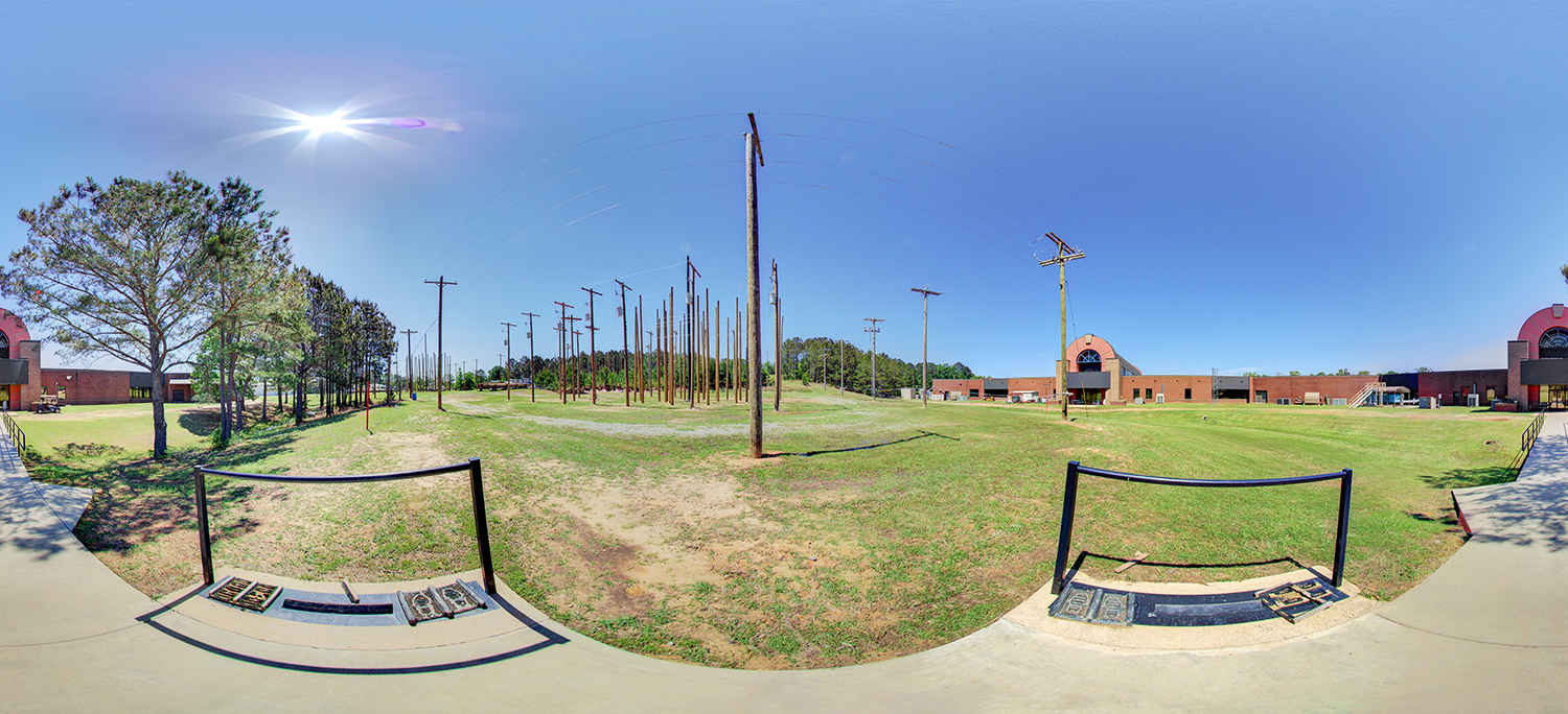 Panoramic view of an outdoor electrical training field with numerous wooden utility poles arranged in rows, grassy open space, nearby campus buildings, and a bright sun overhead in a clear blue sky.