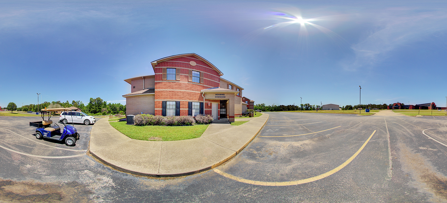 Panoramic view of a two-story brick building labeled “Maintenance Operations,” with a small entrance canopy, surrounded by a wide parking lot, a golf cart, and open campus grounds under a bright sunny sky.