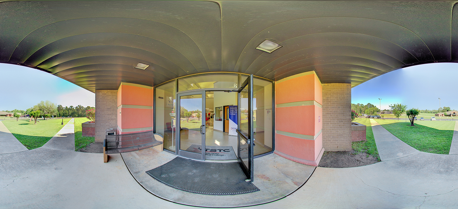 Panoramic view of a campus building entrance with glass double doors propped open beneath a covered overhang, featuring a bench and trash can near the doorway, and walkways leading through a landscaped grassy area on either side.