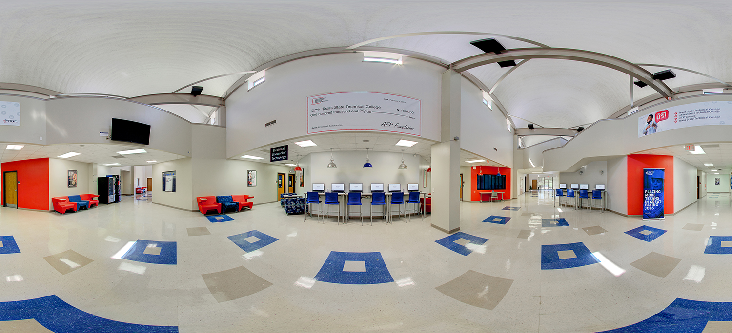 Panoramic view of a bright campus commons area with high curved ceilings, computer workstations, red lounge seating, vending machines, and open hallways connecting academic departments.