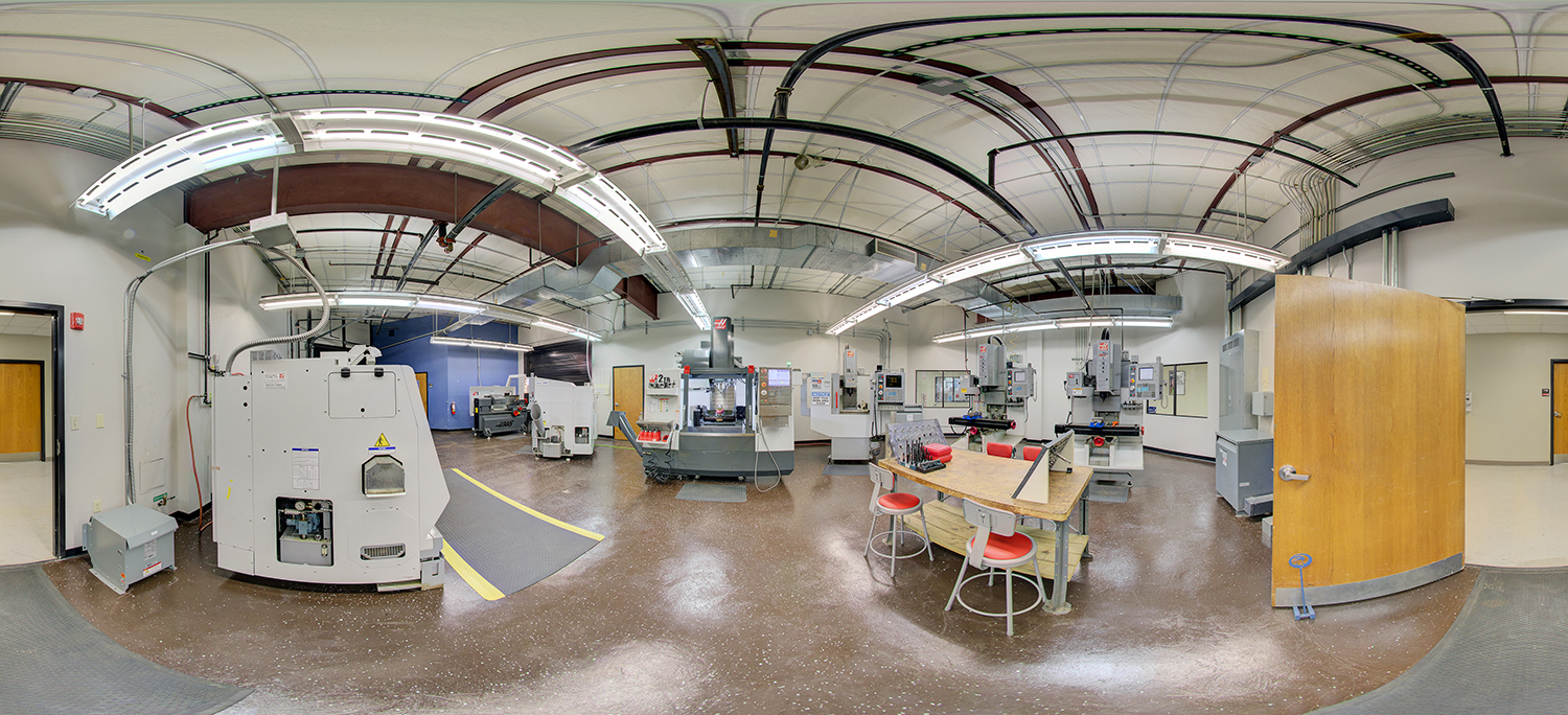 Panoramic view of a machining lab with industrial CNC machines, control panels, and workstations, featuring overhead ductwork, bright fluorescent lighting, and a central table with stools for hands-on training.
