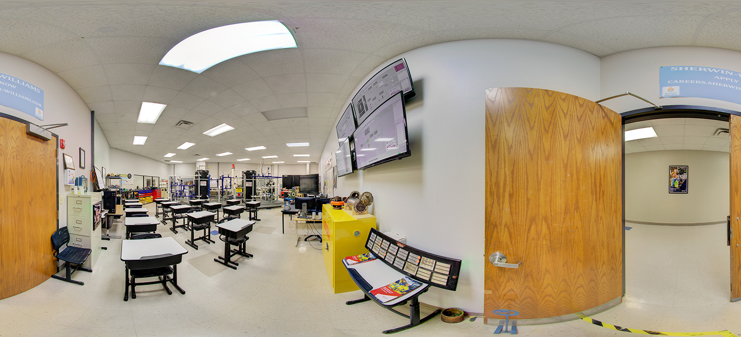 Panoramic view of a classroom with individual student desks facing industrial training equipment, wall-mounted monitors displaying technical content, and open doors leading to adjacent rooms in a lab setting.