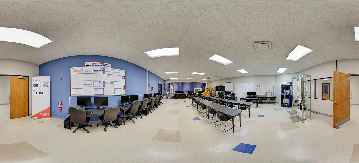 Panoramic view of a technical training classroom with rows of desks and chairs, a line of desktop computers along one wall, instructional posters, and industrial training equipment setups at the back of the room.