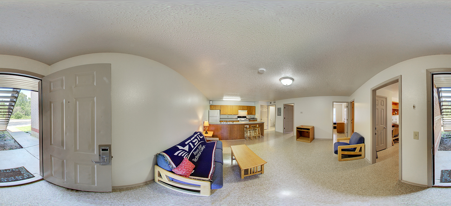 Panoramic view of a modest apartment living area with a small couch, coffee table, chair, and an open kitchen with wooden cabinets and bar stools, connected to bedrooms and an exterior doorway.