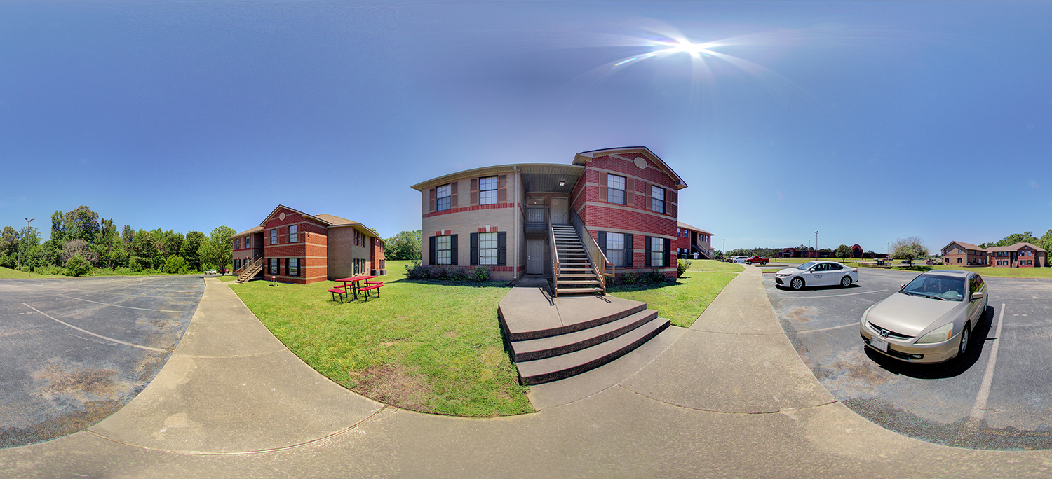 Panoramic view of a small brick apartment-style building with exterior stairs, surrounded by sidewalks, parked cars, grassy areas, and neighboring buildings under a bright midday sun.