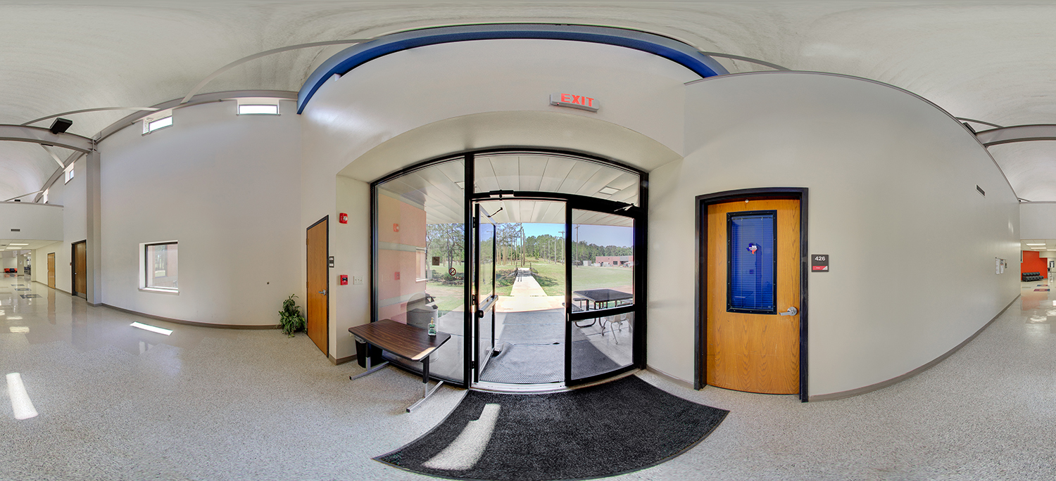 Panoramic view of a building interior entrance with glass double doors leading outside to a walkway and grassy area, an exit sign overhead, nearby hallway doors, and a small table with hand sanitizer beside the entry.