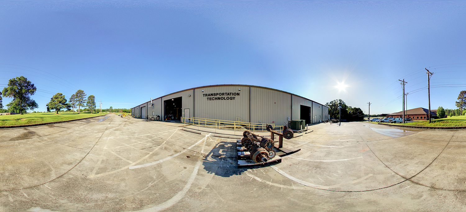 Panoramic exterior view of a large Transportation Technology building with open service bays, surrounded by a wide concrete lot, utility poles, and equipment parts under a bright sunny sky.