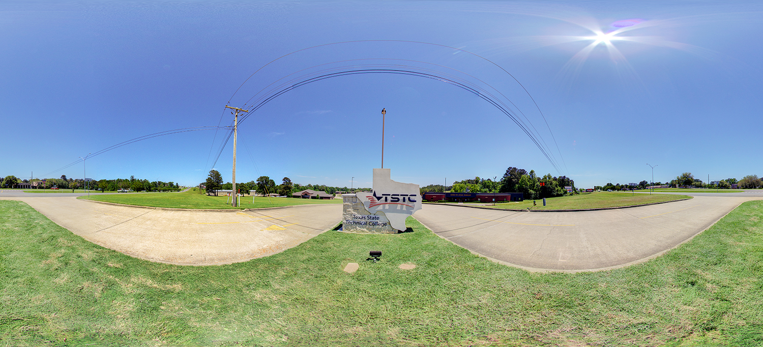 Panoramic view of a Texas State Technical College campus entrance sign set on a grassy median, with surrounding roads, power lines overhead, and low campus buildings in the distance under a clear, sunny sky.