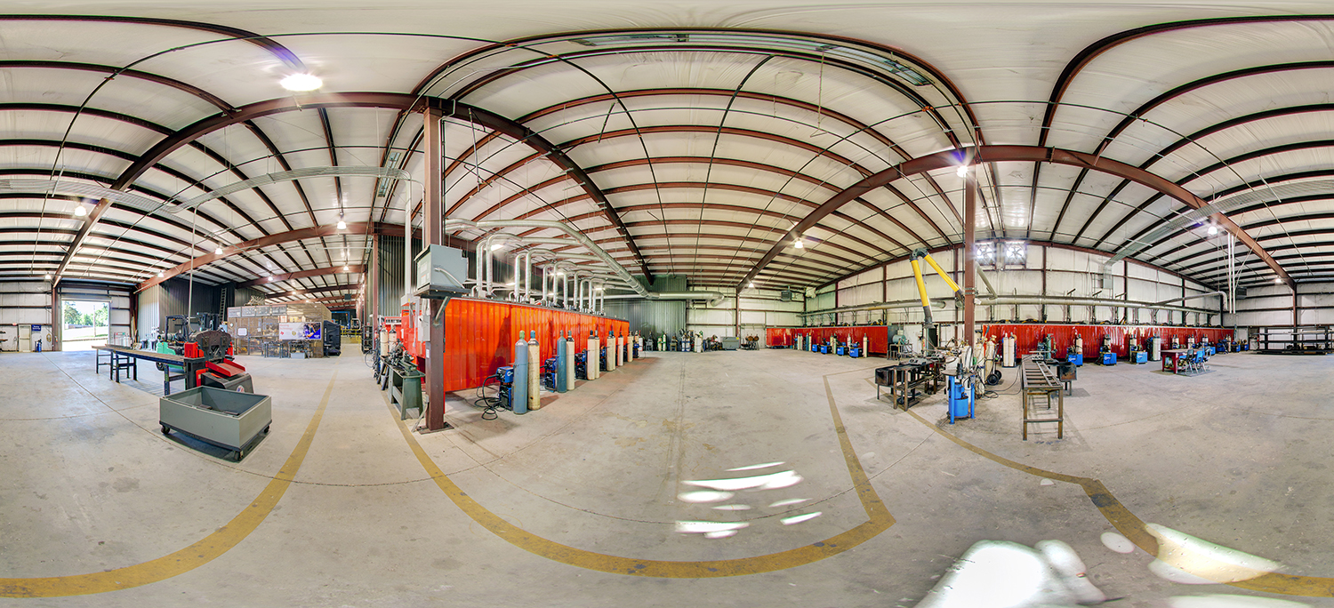 Panoramic view of a spacious welding and fabrication lab with rows of red welding booths, gas cylinders, metal worktables, and ventilation ducts under a high steel-beam ceiling.