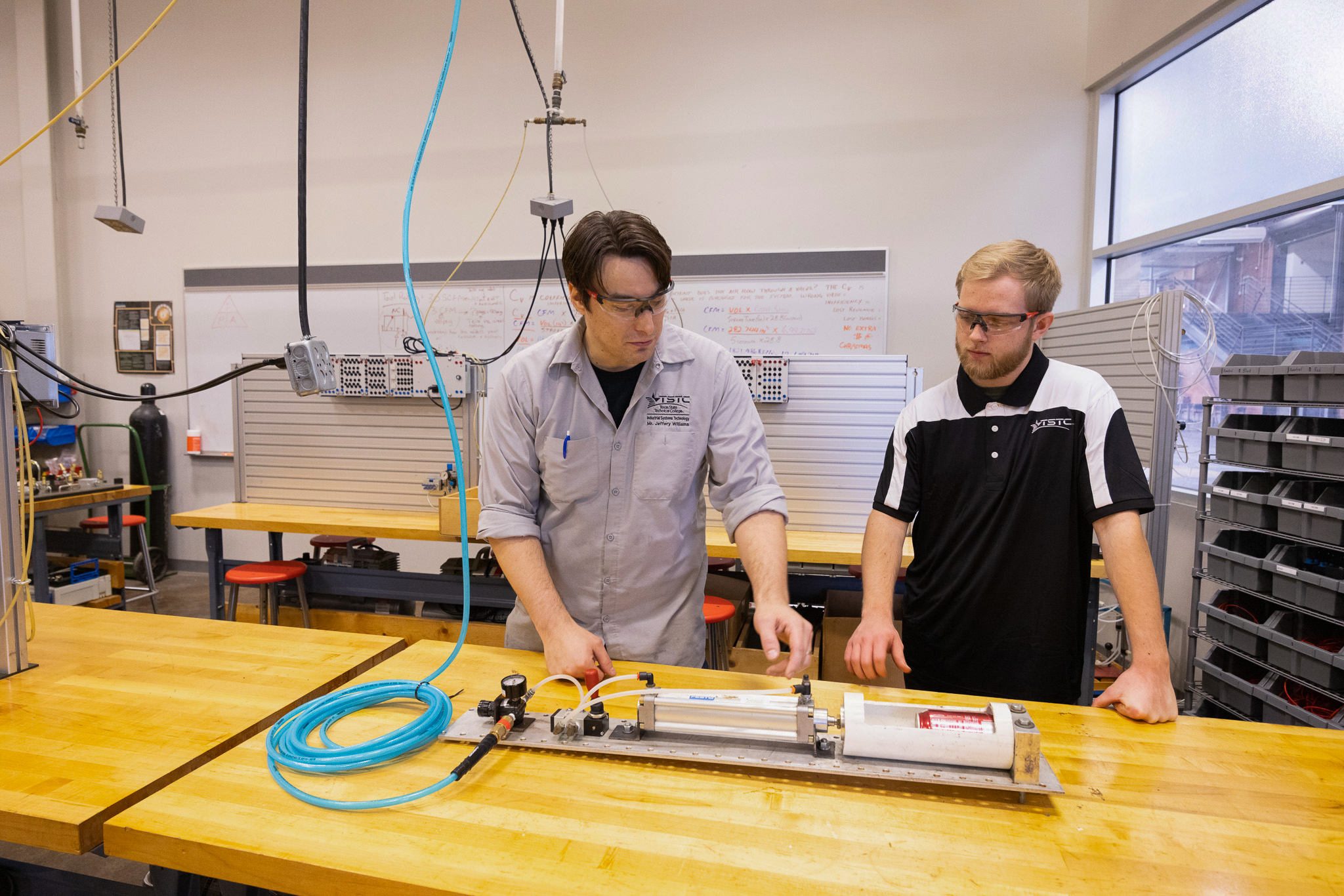 Two students wearing safety glasses stand at a workbench as one points to a pneumatic cylinder setup with hoses and a regulator, explaining its operation while the other observes.