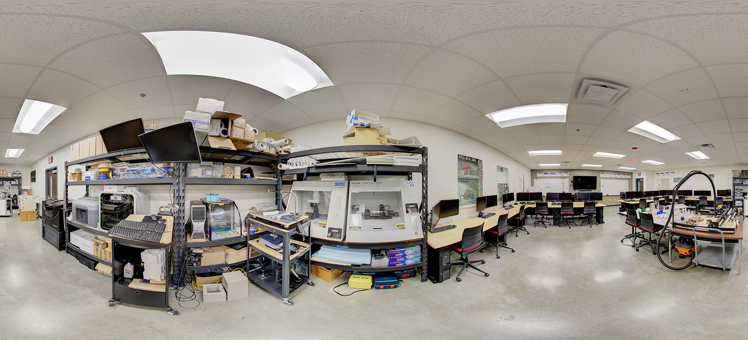 Panoramic view of a technical lab area with metal shelving holding equipment and supplies, various machines and devices, and a row of computer workstations with chairs arranged along the walls for hands-on training.