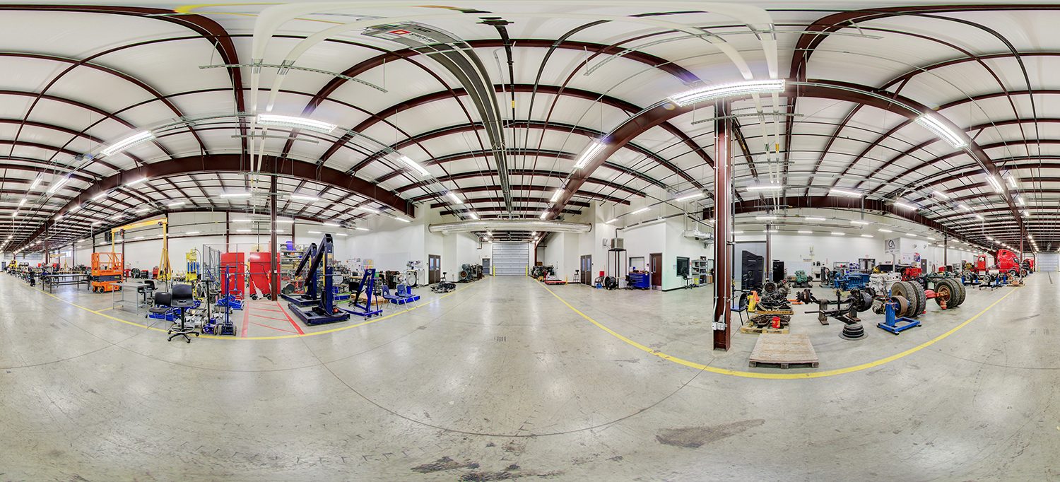 Panoramic view of a large industrial training area with high ceilings, exposed beams, and multiple sections containing mechanical equipment, vehicle components, and workstations arranged across a wide open floor.