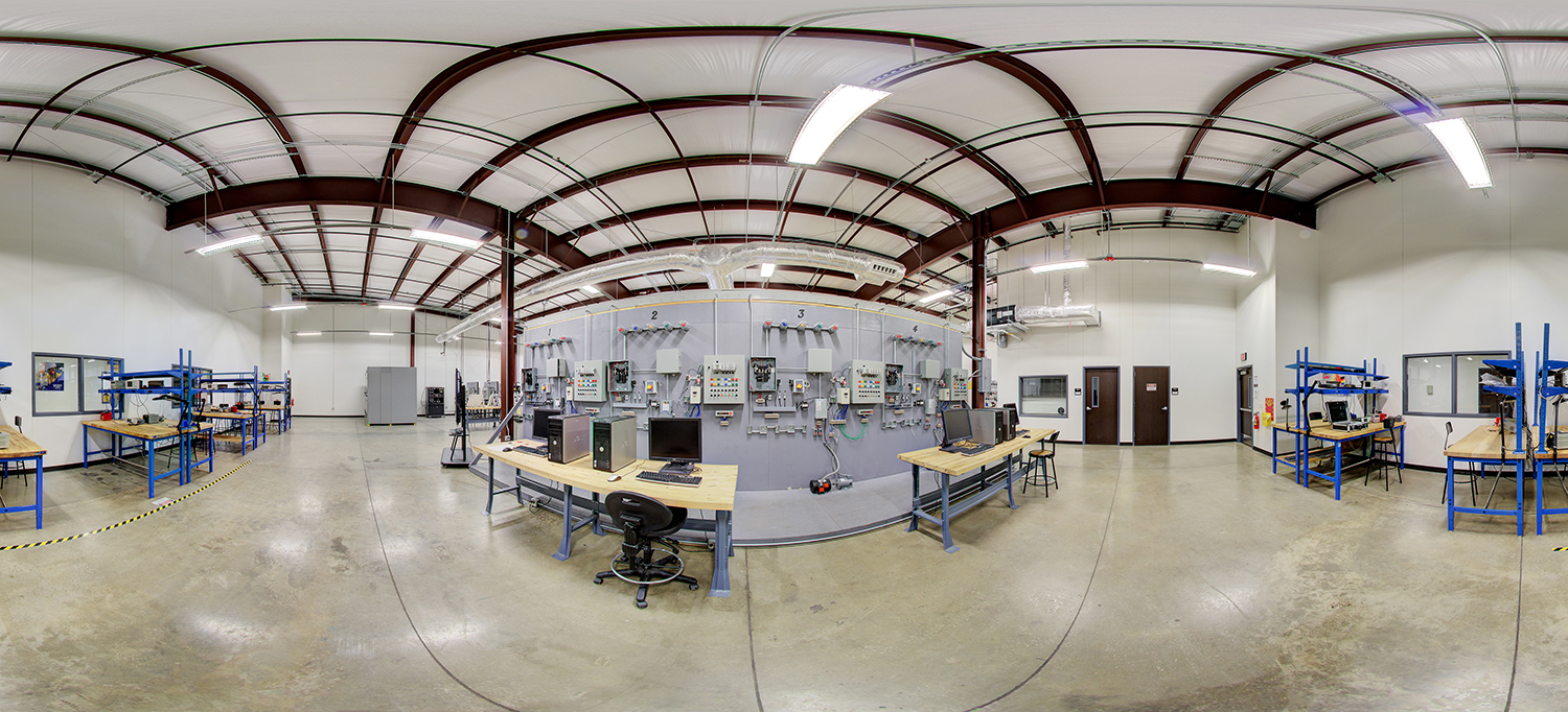 Panoramic view of an electrical training lab area with workbenches, computers, and a central wall of mounted control panels, wiring systems, and industrial electrical components used for hands-on training.