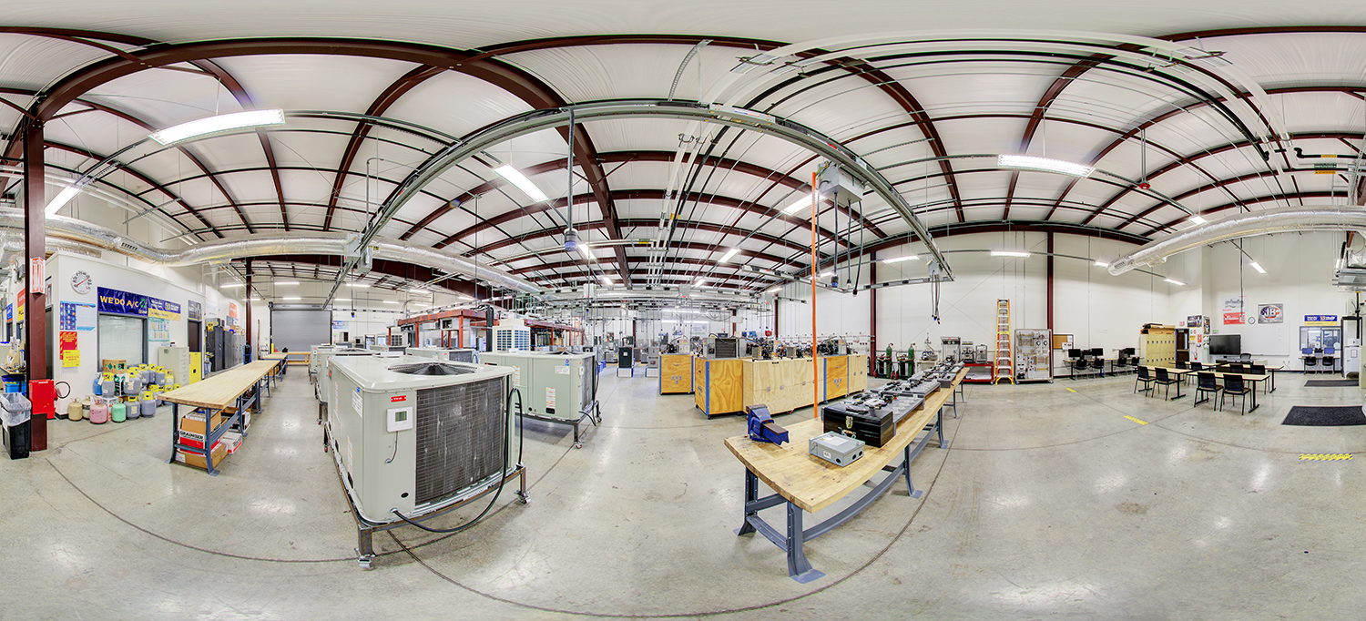 Panoramic view of an HVAC training lab area with large air conditioning units, workbenches with tools and components, overhead piping systems, and open floor space with seating and instructional areas.
