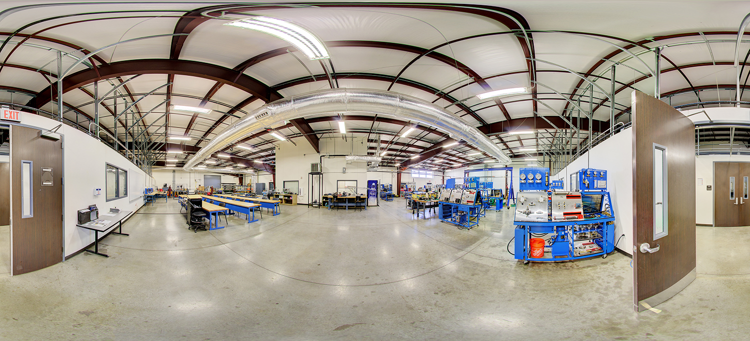 Panoramic view of a technical training lab area with blue industrial workstations, long workbenches, tools and equipment, overhead ductwork and piping, and wide open floor space with multiple entry doors.