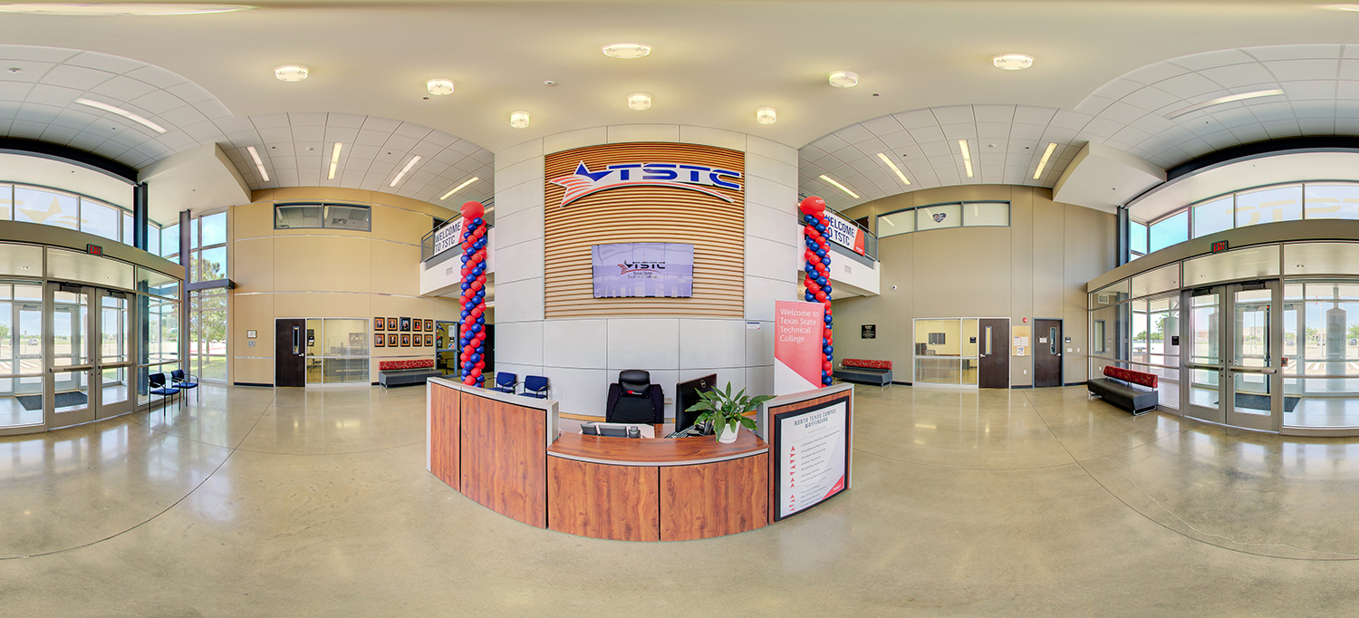 Panoramic view of a campus lobby area with a central reception desk, TSTC signage on the wall behind it, red and blue balloon columns, seating along the walls, and multiple glass entry doors on both sides.