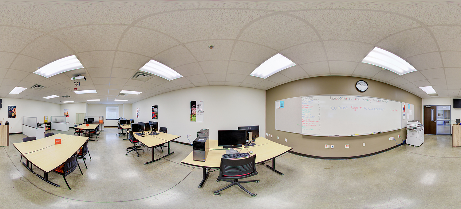 Panoramic view of a computer lab area with multiple desks, desktop computers, office chairs, a large whiteboard with writing, posters on the walls, and printers along the side.
